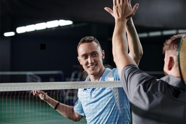 Zwei Männer reichen sich fröhlich die Hand über ein Badminton-Netz in einer Sporthalle.