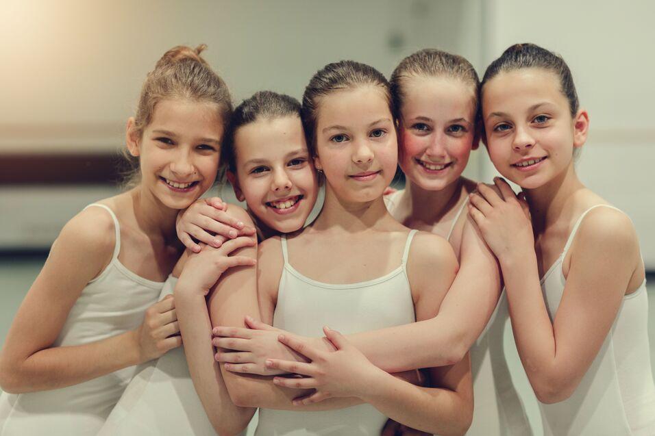 Fünf junge Balletttänzerinnen in weißen Kostümen lächeln und umarmen sich, vor einem hellen Hintergrund.