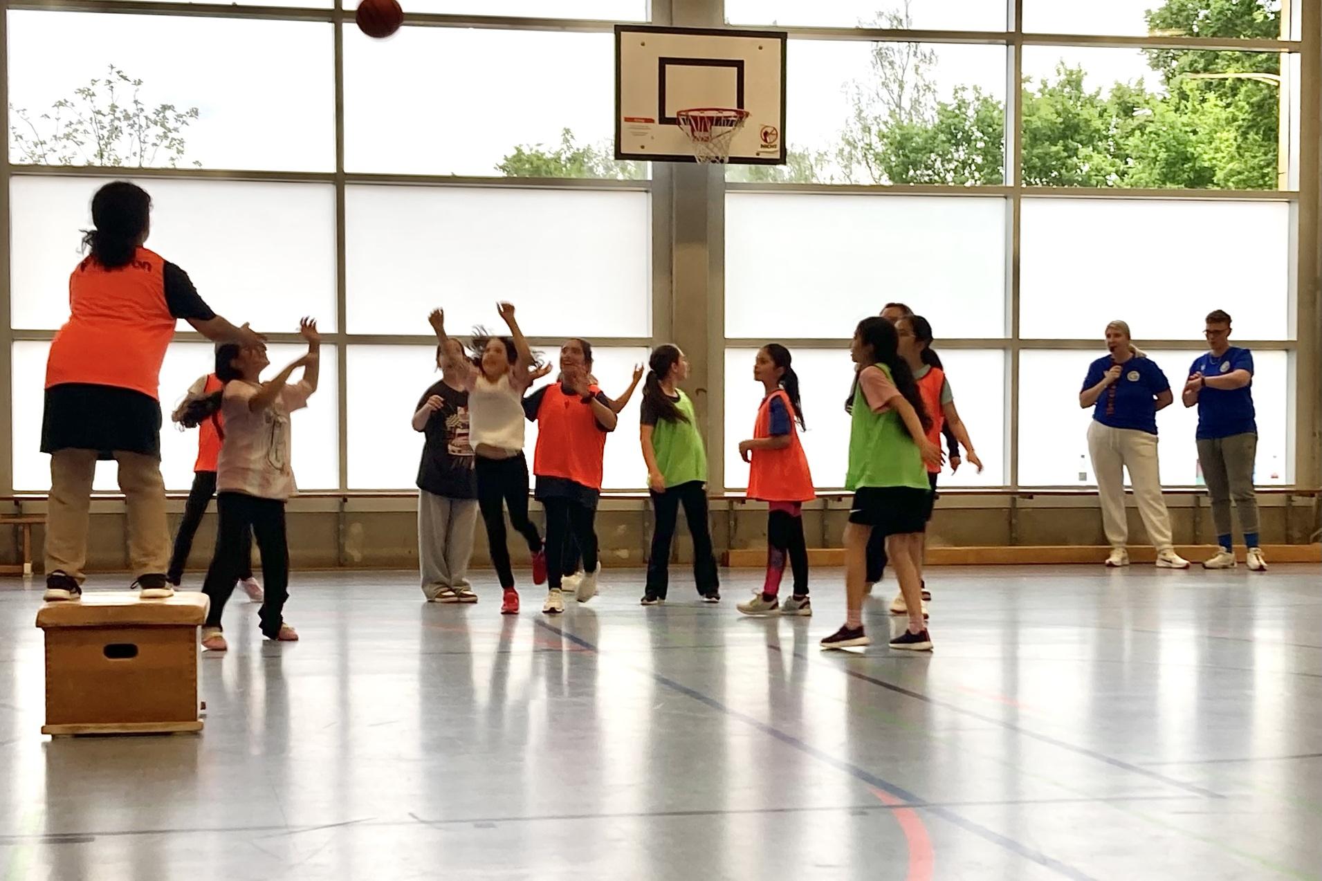 Gruppe von Kindern in Sportkleidung spielt Basketball in einer Halle, während Trainer die Übungen anleiten.