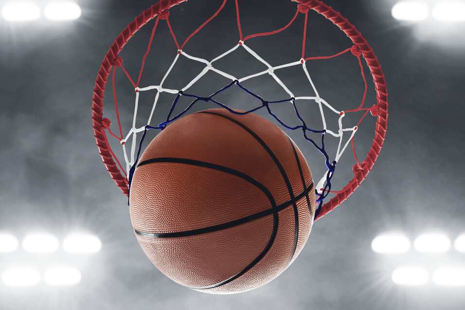 Nahaufnahme eines Basketballs, der durch den Basketballkorb in einem beleuchteten Sportumfeld fliegt.