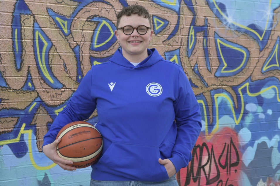 Person in blauer Sportjacke mit Brille h&auml;lt einen Basketball vor einer bunten Graffiti-Wand.