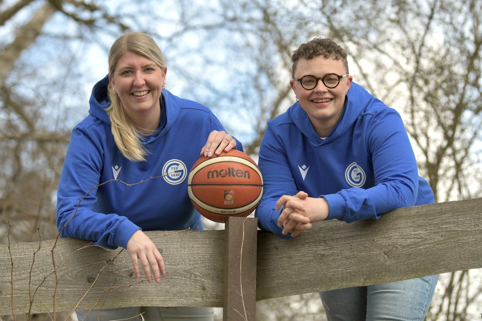 Zwei Frauen in blauen Hoodies stehen an einem Holzgel&auml;nder und halten einen Basketball. Hintergrund mit B&auml;umen und Himmel.