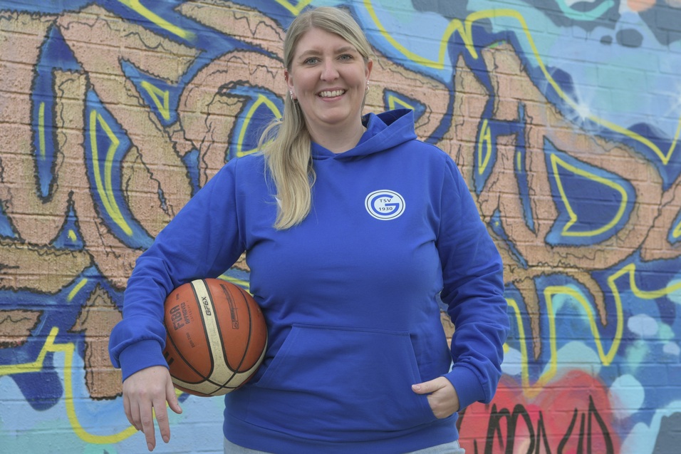 Frau in blauer Kapuzenjacke mit Basketball vor einer bunten Graffiti-Wand.