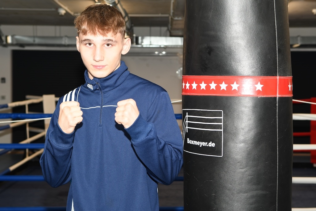 Junger Boxer steht vor einem Boxsack in einem Trainingsraum und zeigt eine Kampfposition.