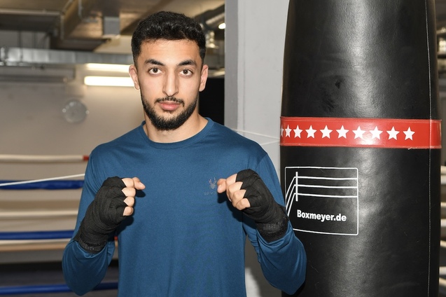 Junger Boxer steht vor einem Boxsack und hat die F&auml;uste zum Kampfstil erhoben, in einer Sporthalle.