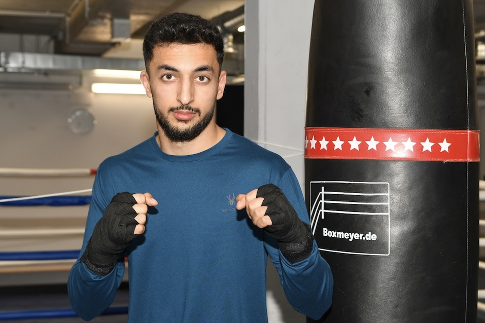 Junger Boxer steht vor einem Boxsack und hat die F&auml;uste zum Kampfstil erhoben, in einer Sporthalle.