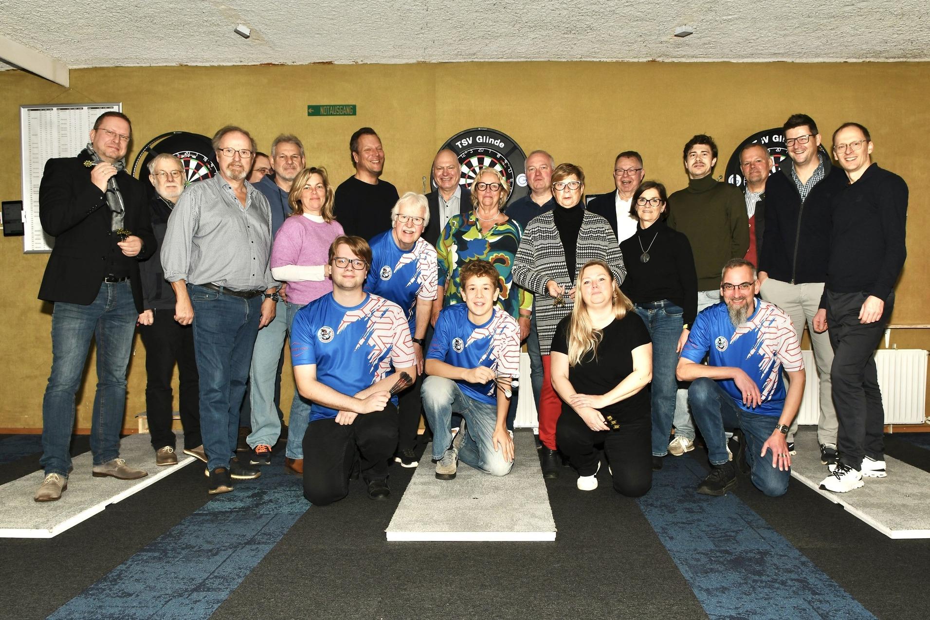 Gruppenscreenshot von 18 Personen in einem Dartsalon, einige tragen Dartshirts, Hintergrund mit Dartscheiben.