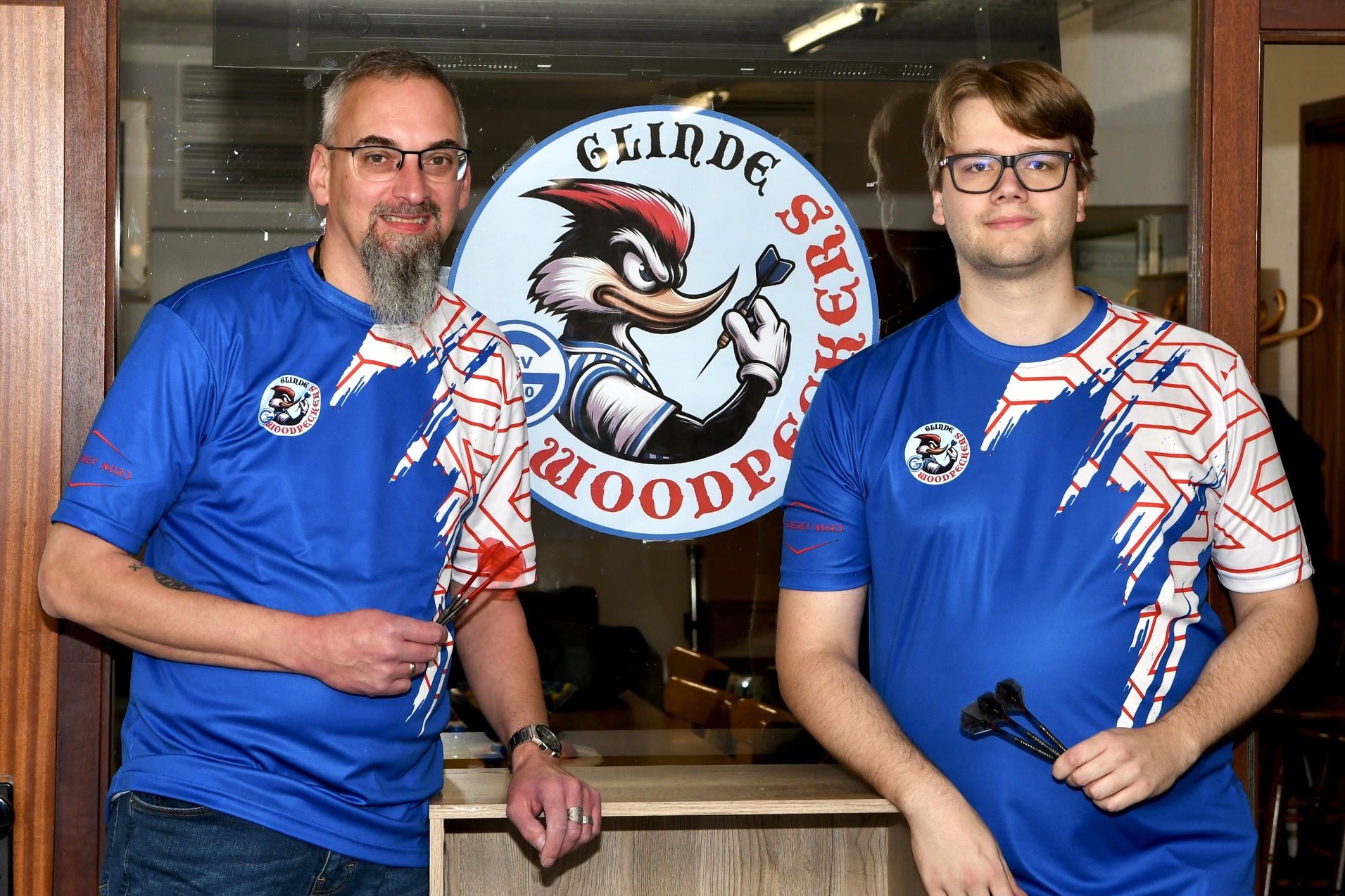 Zwei M&auml;nner in blauen Sporttrikots stehen vor einem Logo mit dem Schriftzug &bdquo;Glinde Woodpockers&ldquo;.