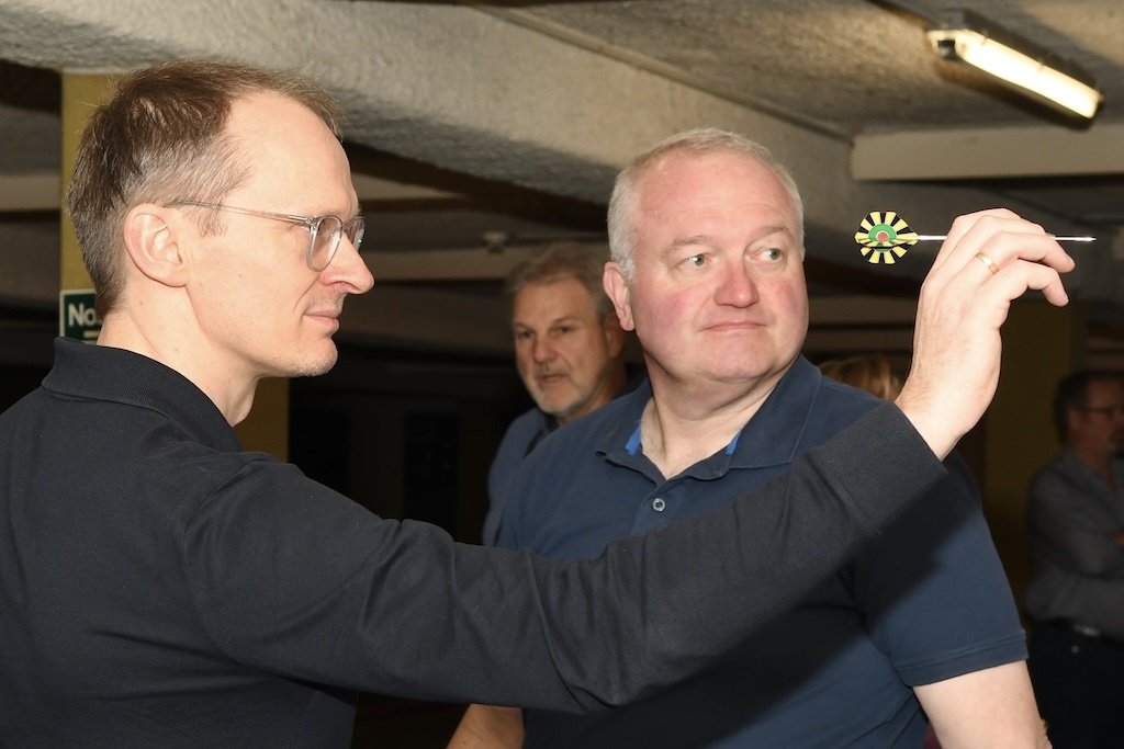 Zwei M&auml;nner stehen in einem Dartsalon; einer zielt mit einem Dart auf die Dartscheibe, der andere beobachtet gespannt.