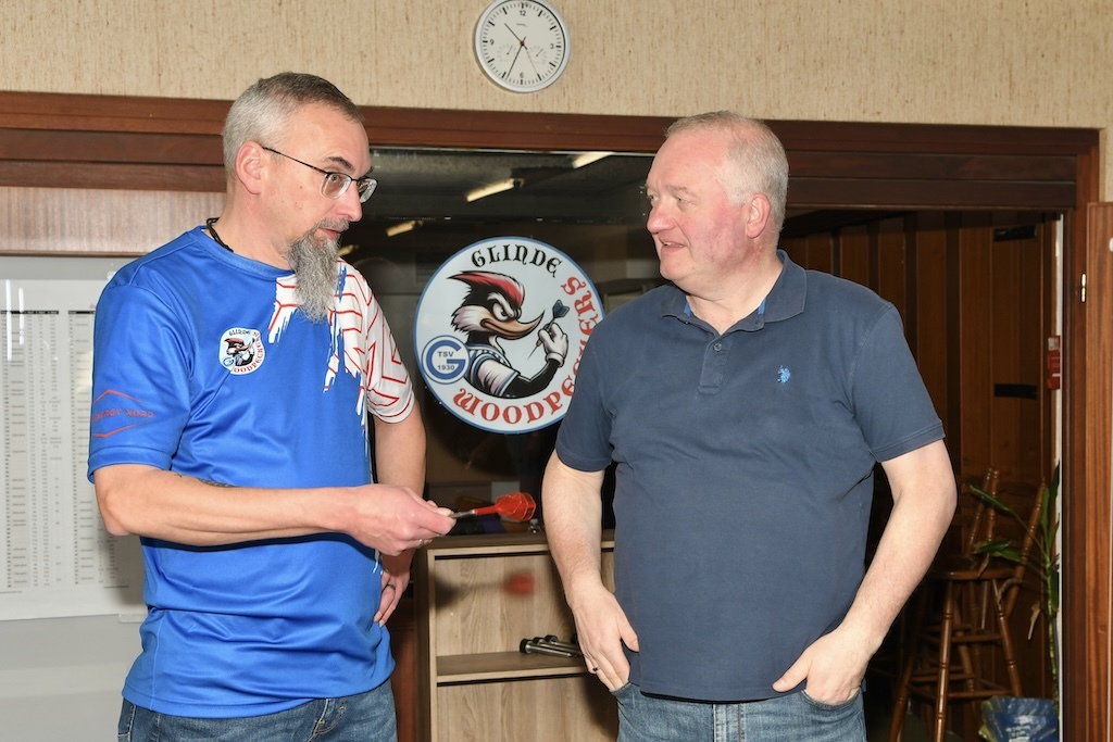 Zwei M&auml;nner im Gespr&auml;ch, einer h&auml;lt eine Dartscheibe, im Hintergrund eine Uhr und ein Logo an der Wand.