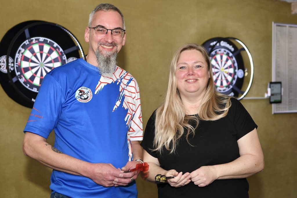 Zwei Personen stehen l&auml;chelnd vor Dartboards, einer tr&auml;gt ein blaues Shirt, die andere ein schwarzes T-Shirt.