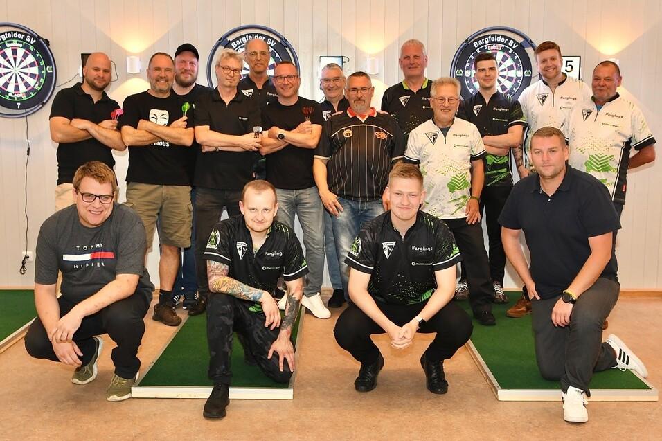 Gruppe von 15 Dartspielern in sportlichen Outfits posiert vor Dartboards auf einer Turnierfl&auml;che.