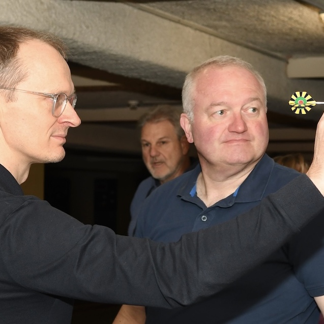 Zwei M&auml;nner stehen in einem Dartsalon; einer zielt mit einem Dart auf die Dartscheibe, der andere beobachtet gespannt.