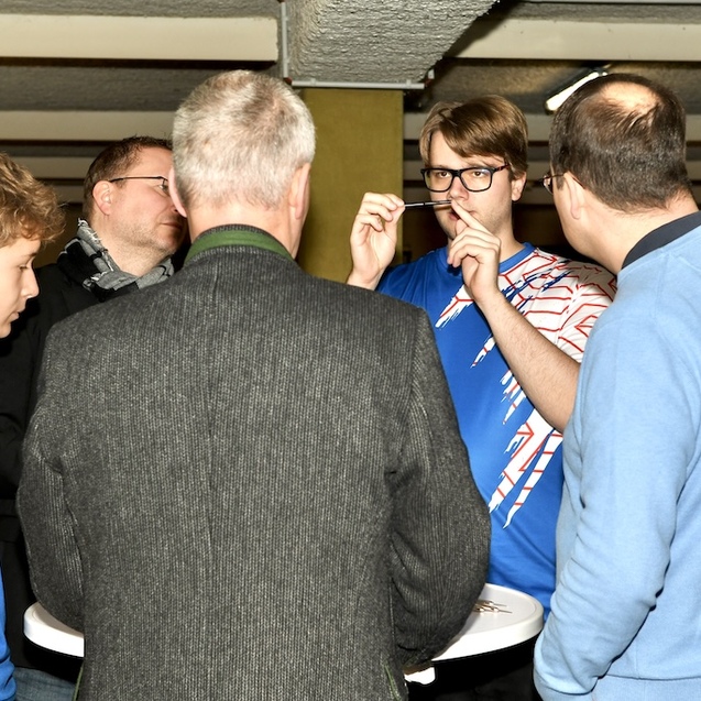 Gruppe von Menschen diskutiert an einem Stehtisch, w&auml;hrend ein junger Mann mit Brille gestikuliert.