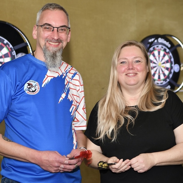 Zwei Personen stehen l&auml;chelnd vor Dartboards, einer tr&auml;gt ein blaues Shirt, die andere ein schwarzes T-Shirt.