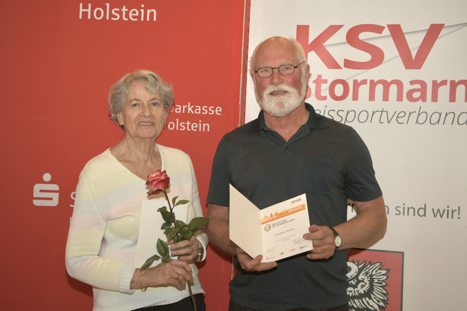 Eine &auml;ltere Frau mit Rose &uuml;berreicht einem Mann mit Brille eine Urkunde vor einem KSV Stormarn Banner.