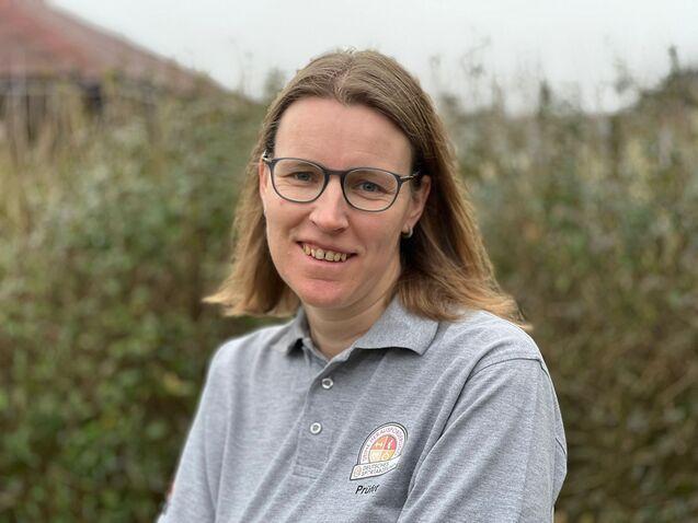 Frau mit Brille und blonder Haar trägt ein graues Polo-Shirt, steht vor einer natürlichen, grünen Hintergrundlandschaft.