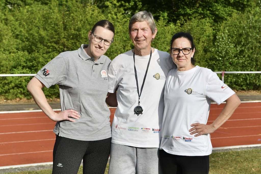 Drei Personen posieren auf einer Laufbahn, zwei Frauen in Sportoutfits, ein Mann mit Trainerpfeife zwischen ihnen.