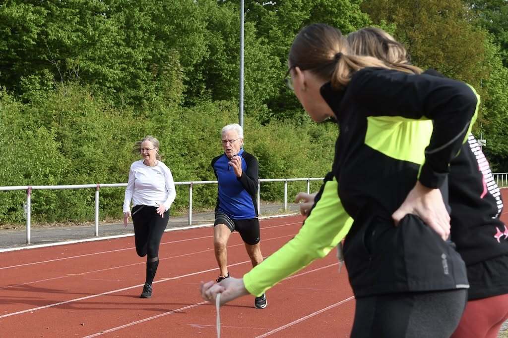 Vier Sportler laufen auf einer Laufbahn, w&auml;hrend eine Person die Zeit stoppt. Gr&uuml;nfl&auml;chen im Hintergrund.