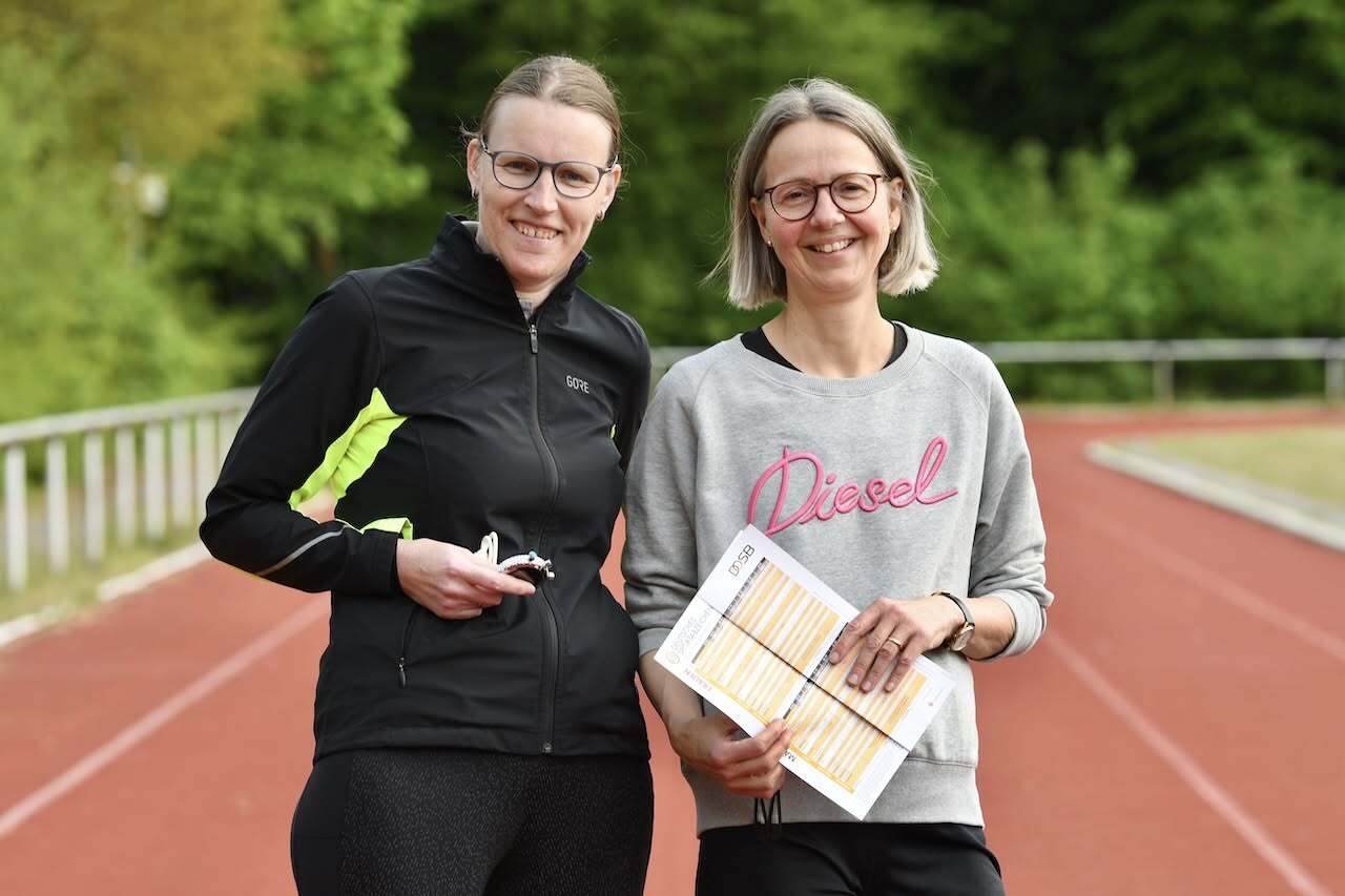 Zwei Frauen stehen auf einer Laufbahn, eine hält Notizen, die andere hat einen Schlüsselbund in der Hand.