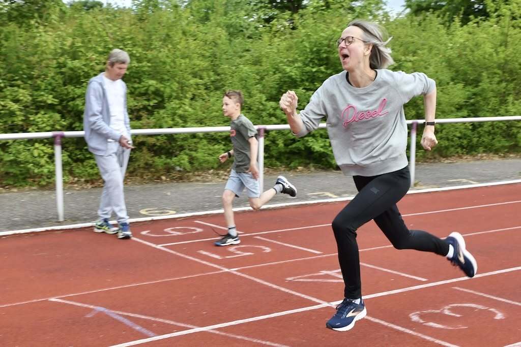 Frau in Sportkleidung läuft sprintend auf der Bahn, neben ihr zwei Kinder in Bewegung. Grüne Büsche im Hintergrund.