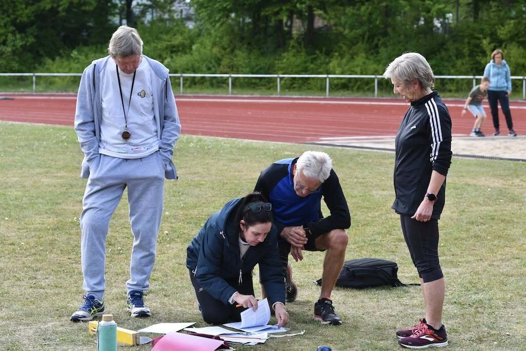 Vier Personen auf einem Sportplatz, eine kniet mit Unterlagen, die anderen beobachten und diskutieren.