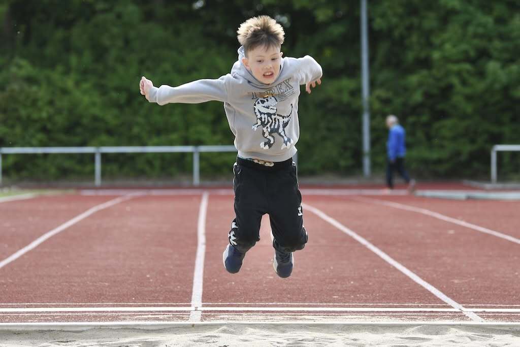 Junge springt fröhlich in die Weitsprunggrube auf einer roten Tartan-Leichtathletikanlage.