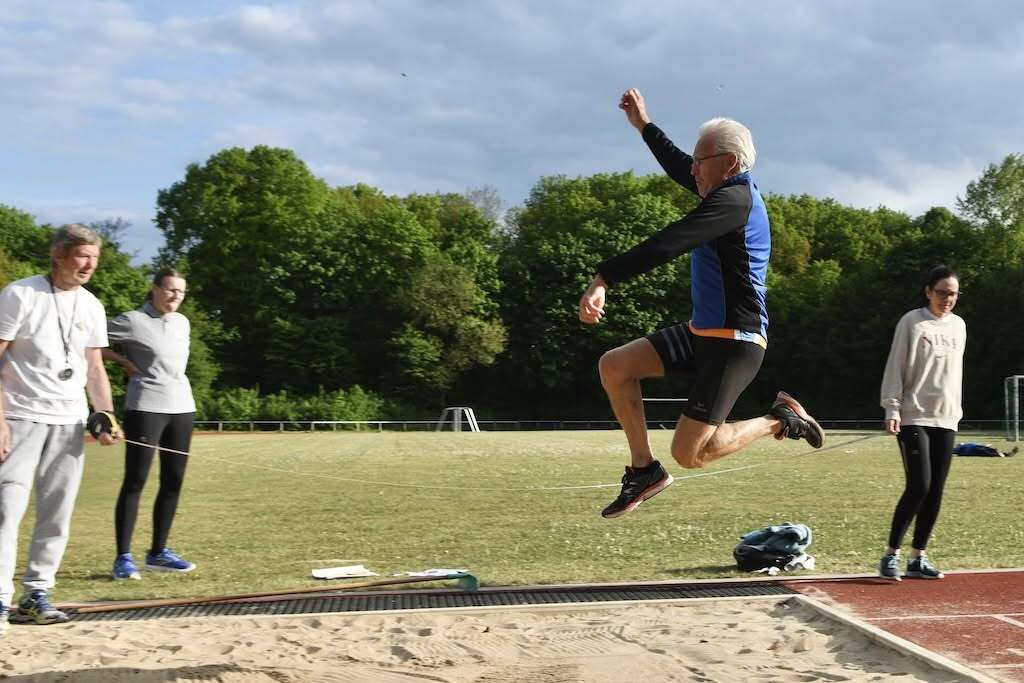 Mann springt vom Anlaufbrett in die Sandgrube, während Zuschauer auf einem Sportplatz zu sehen sind.