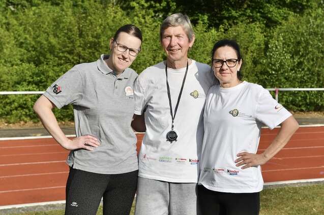 Drei Personen posieren auf einer Laufbahn, zwei Frauen in Sportoutfits, ein Mann mit Trainerpfeife zwischen ihnen.