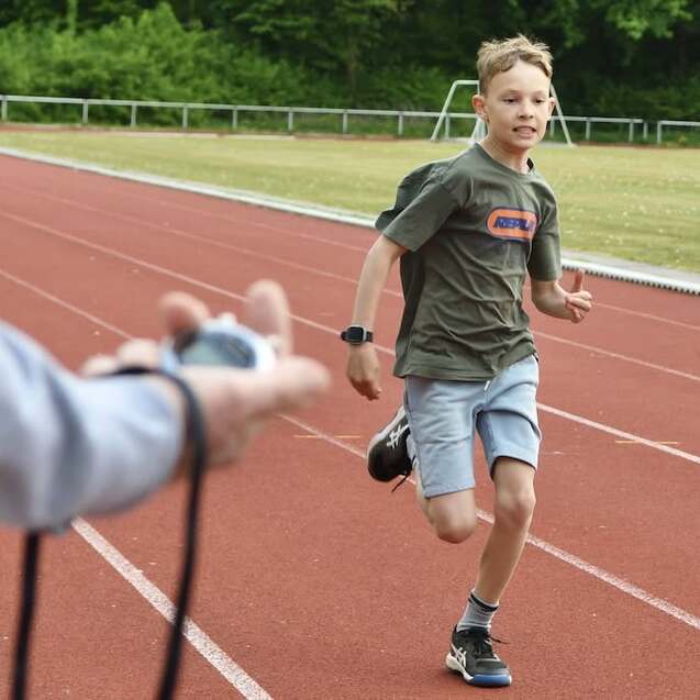 Junge läuft auf einer Laufbahn, während eine Person mit Stoppuhr die Zeit misst. Grüne Landschaft im Hintergrund.