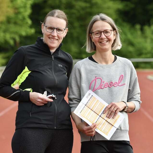 Zwei Frauen stehen auf einer Laufbahn, eine hält Notizen, die andere hat einen Schlüsselbund in der Hand.