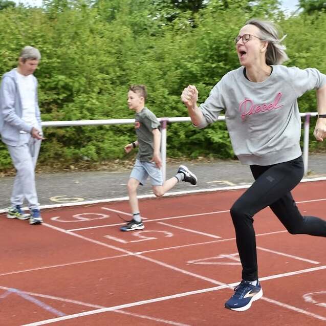 Frau in Sportkleidung läuft sprintend auf der Bahn, neben ihr zwei Kinder in Bewegung. Grüne Büsche im Hintergrund.