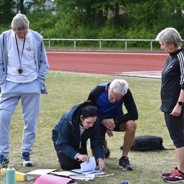 Vier Personen auf einem Sportplatz, eine kniet mit Unterlagen, die anderen beobachten und diskutieren.