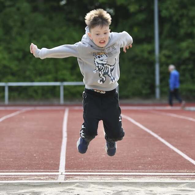 Junge springt fröhlich in die Weitsprunggrube auf einer roten Tartan-Leichtathletikanlage.