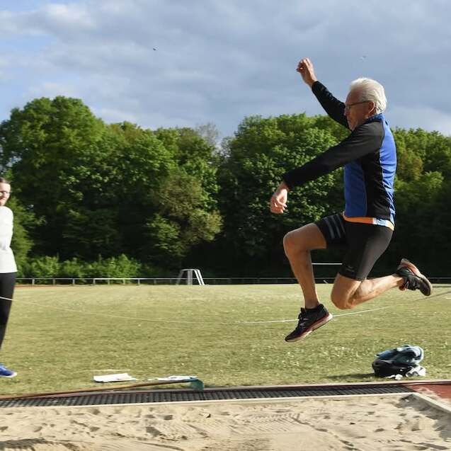 Mann springt vom Anlaufbrett in die Sandgrube, während Zuschauer auf einem Sportplatz zu sehen sind.