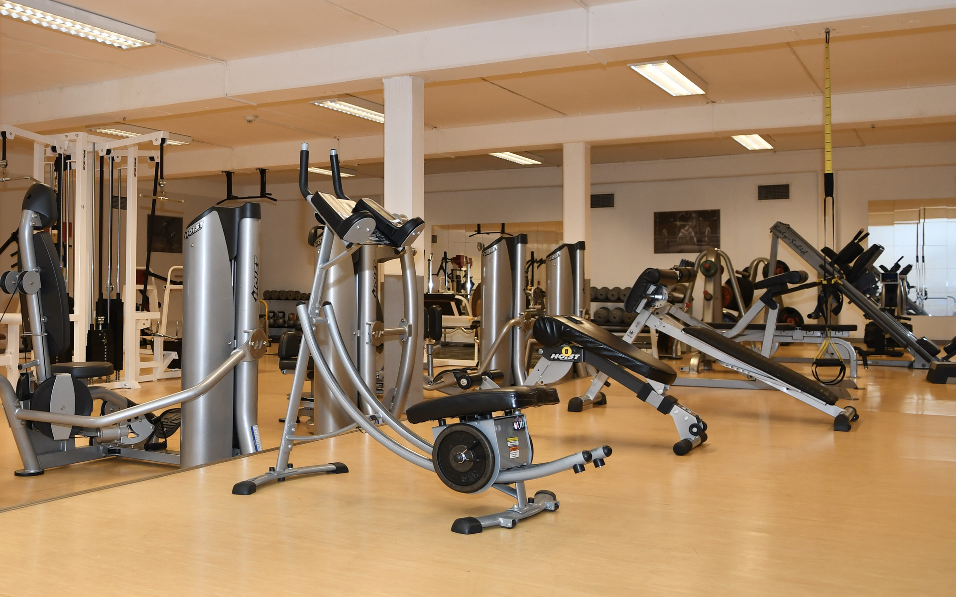 Fitnessraum mit verschiedenen Trainingsger&auml;ten, darunter Laufband, Ruderger&auml;t und Kraftstationen auf Holzfu&szlig;boden.