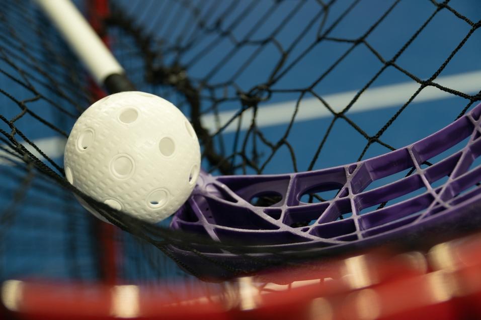 Weißer Plastikball liegt in einem violetten Netz von einem Hockeyschläger auf einem blauen Sportboden.