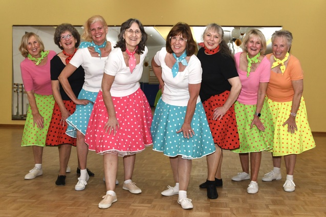 Gruppe von acht Frauen in bunten Polka-Dot-R&ouml;cken und T-Shirts in einer Tanzpose in einem Saal.