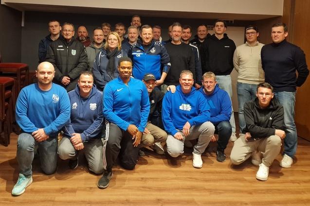 Gruppenfoto von Sportvereinsmitgliedern in blauen und schwarzen Outfits, auf einem Holzfu&szlig;boden zusammengestellt.