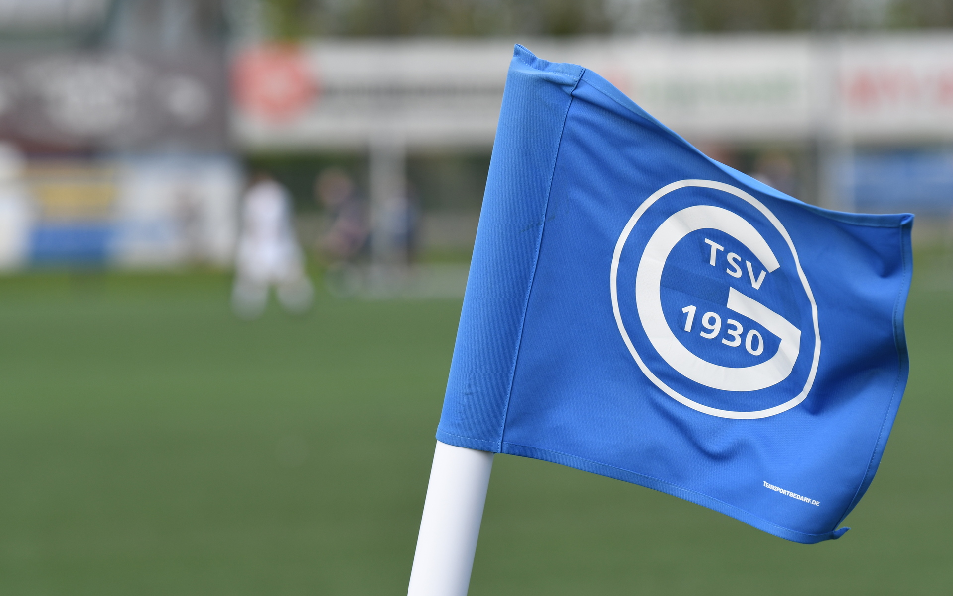 Nahaufnahme einer blauen Eckfahne mit dem Logo des TSV 1930 auf einem Fu&szlig;ballfeld im Hintergrund.