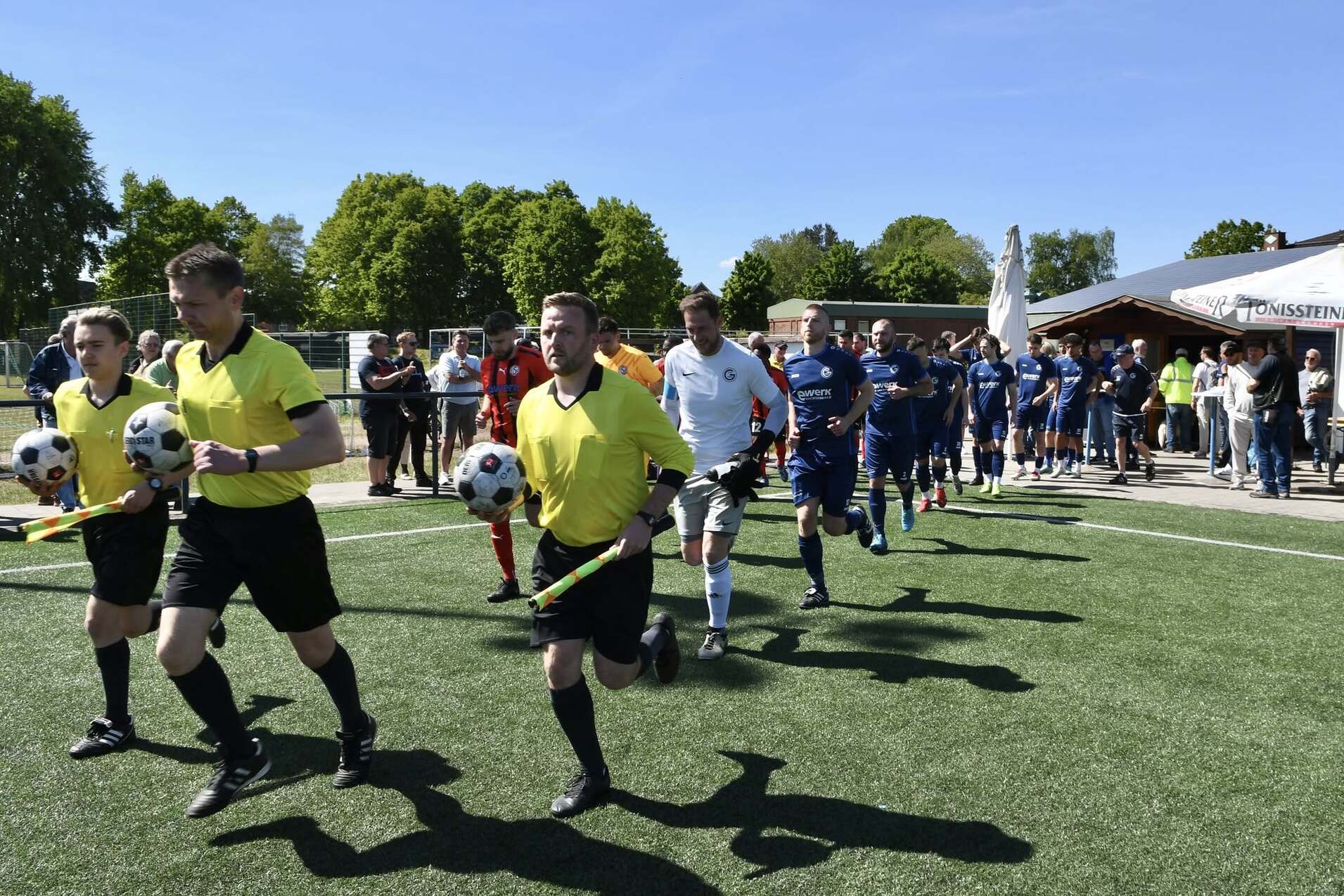 Spieler und Schiedsrichter betreten ein Fußballfeld, während Zuschauer an der Seite stehen und eine sonnige Atmosphäre genießen.