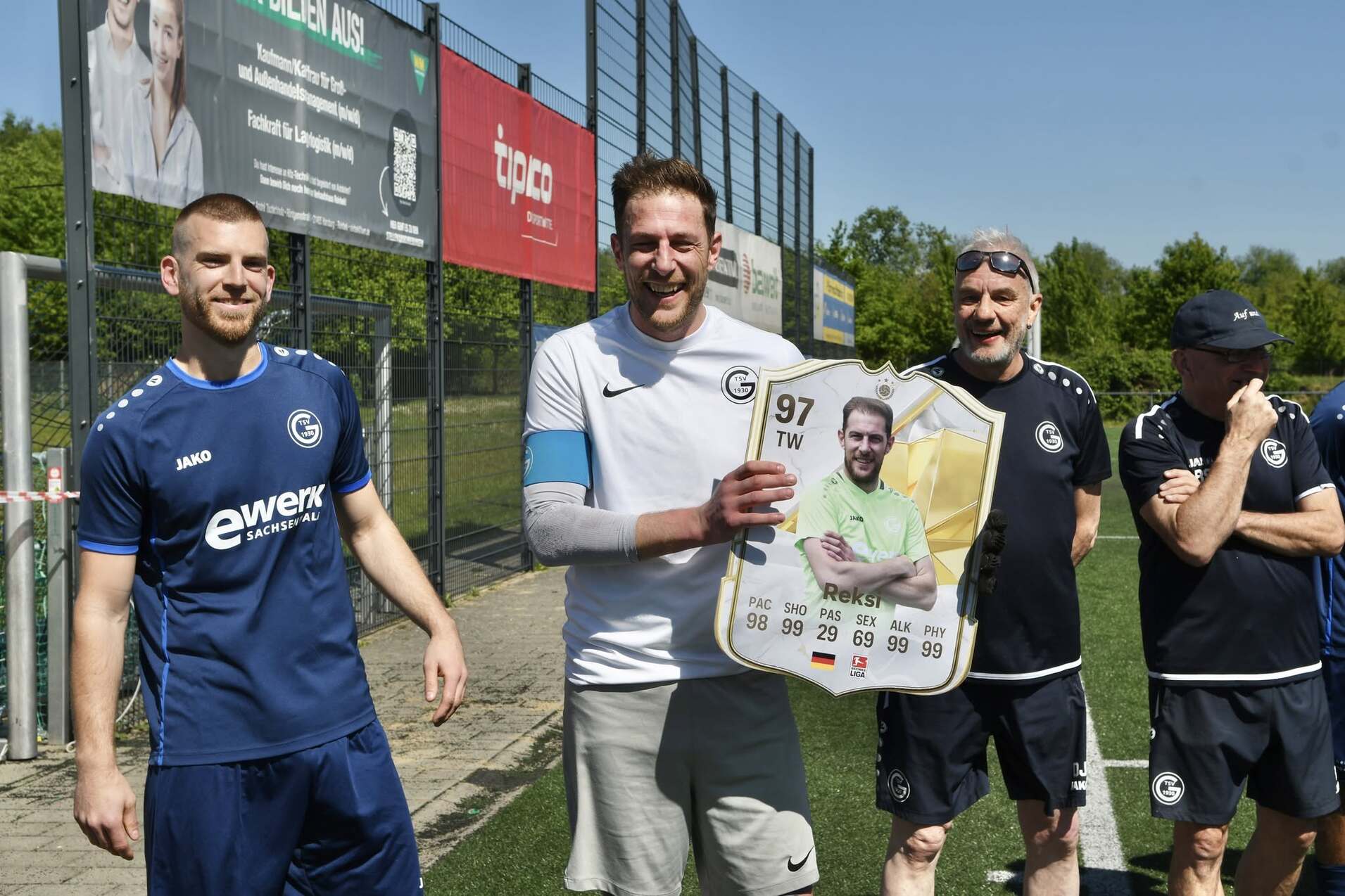 Vier Fußballspieler stehen auf einem Platz, einer hält ein großes Fußballkarten-Poster in den Händen.