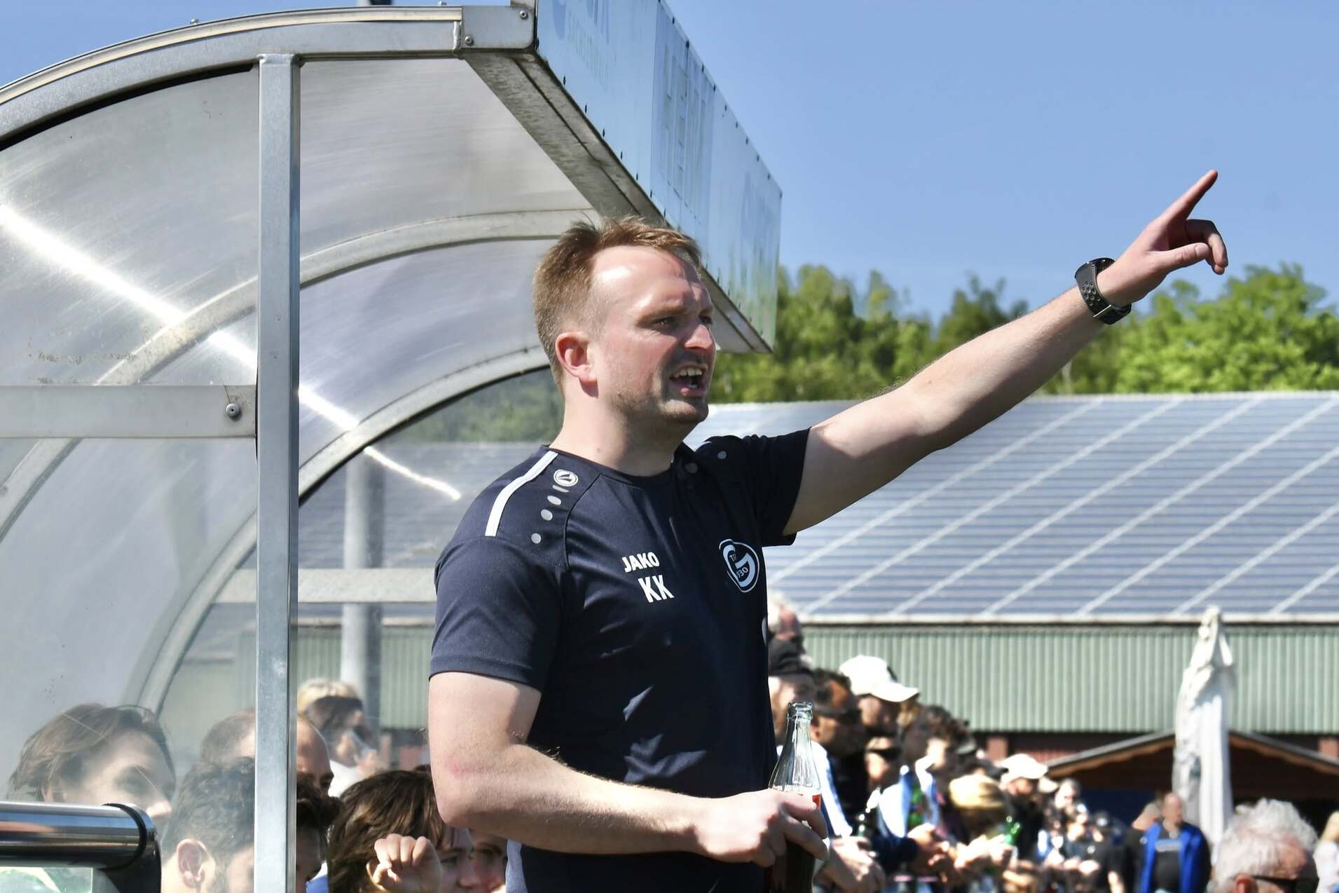 Trainer gibt Anweisungen während des Spiels, im Hintergrund stehen Zuschauer auf einem Sportgelände.