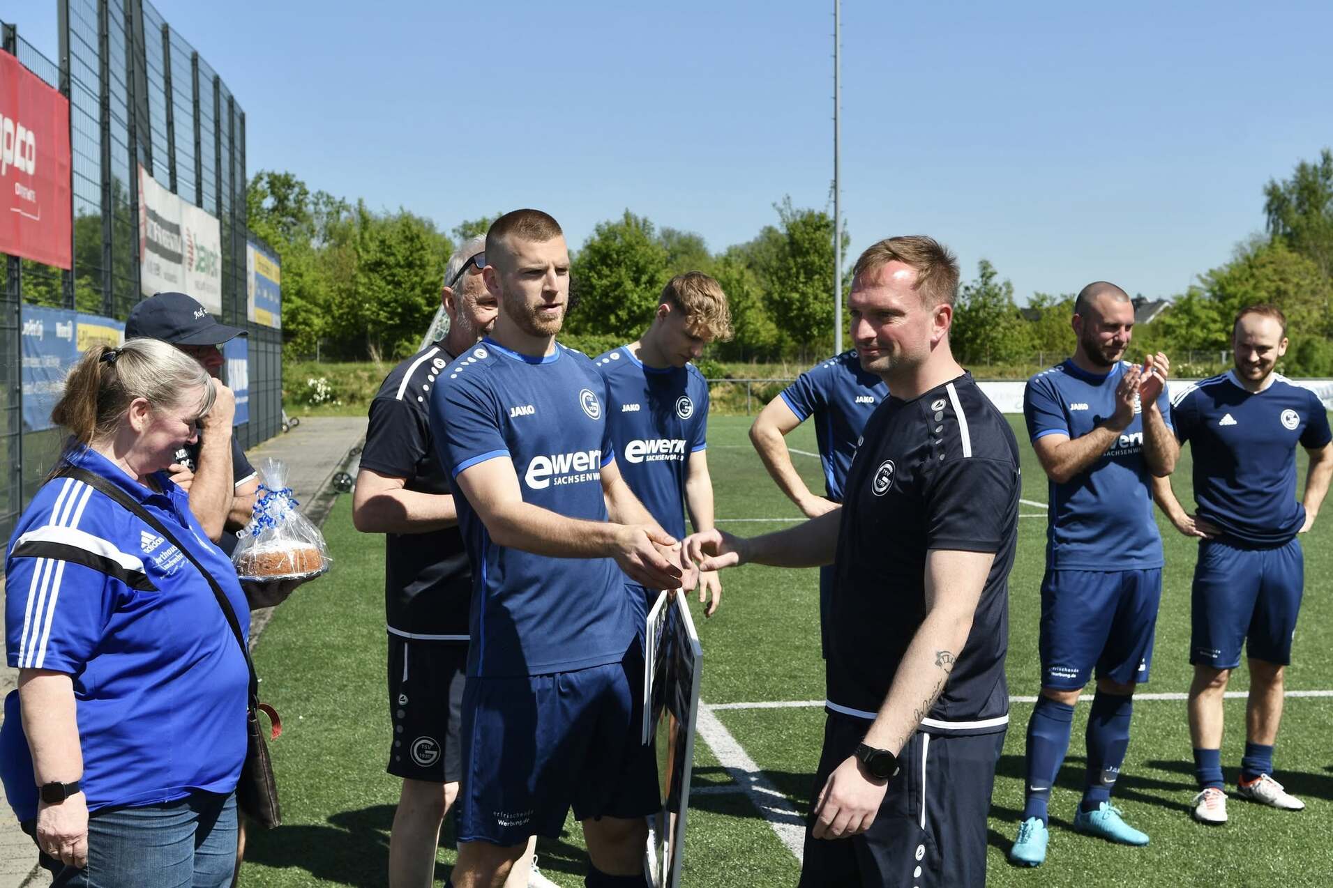 Zwei Fußballspieler übergeben sich eine Auszeichnung auf einem Sportplatz, im Hintergrund applaudierende Teamkollegen.
