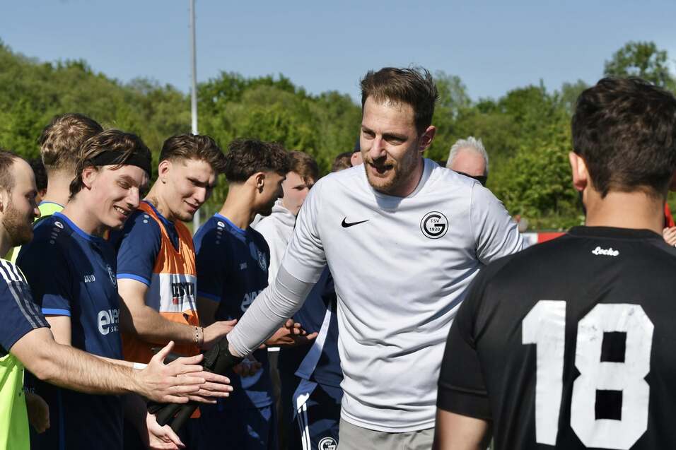 Trainer sch&uuml;ttelt Spielern die Hand auf einem Fu&szlig;ballplatz bei sonnigem Wetter, im Hintergrund B&auml;ume und Zuschauer.