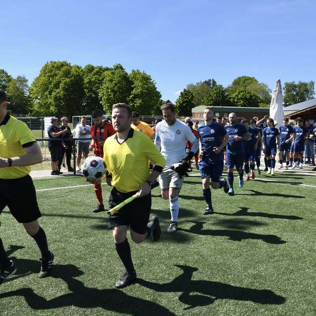 Spieler und Schiedsrichter betreten ein Fußballfeld, während Zuschauer an der Seite stehen und eine sonnige Atmosphäre genießen.