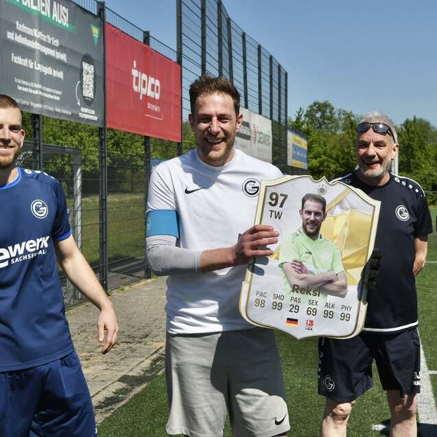 Vier Fußballspieler stehen auf einem Platz, einer hält ein großes Fußballkarten-Poster in den Händen.