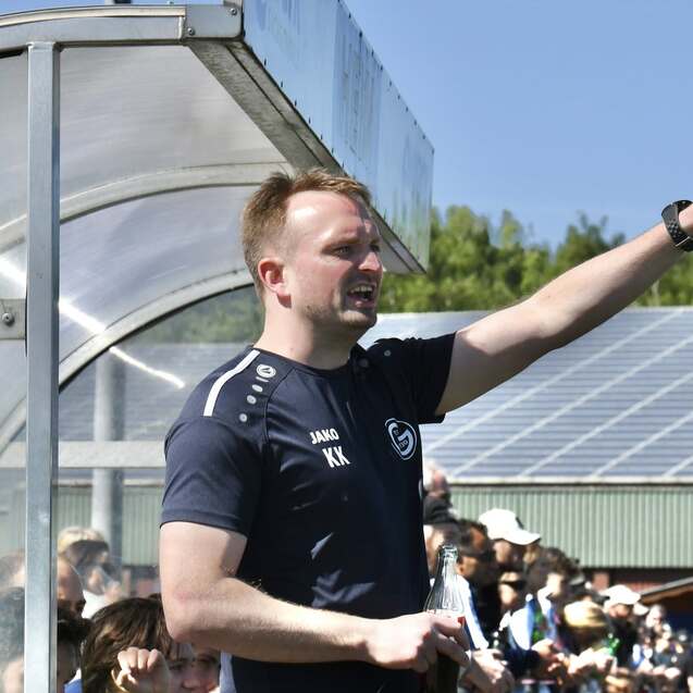 Trainer gibt Anweisungen während des Spiels, im Hintergrund stehen Zuschauer auf einem Sportgelände.