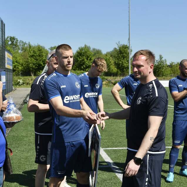 Zwei Fußballspieler übergeben sich eine Auszeichnung auf einem Sportplatz, im Hintergrund applaudierende Teamkollegen.