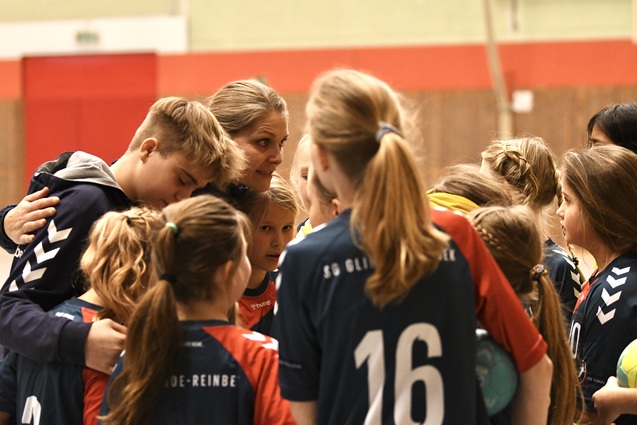 Trainerin spricht mit einer Gruppe von M&auml;dchen in Handballtrikots vor einem Sportplatz. Einige h&ouml;ren aufmerksam zu.